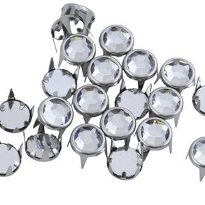 May include: A collection of silver metal studs with clear, faceted, round crystal centres. Each stud has a pointed base and a decorative, cat-ear-shaped top. The studs are scattered against a white background.
