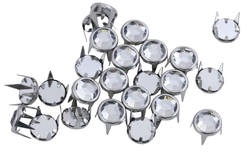 May include: A collection of silver metal studs with clear, faceted, round crystal centres. Each stud has a pointed base and a decorative, cat-ear-shaped top. The studs are scattered against a white background.