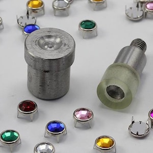 May include: An assortment of colorful rhinestone rivets and setting tools. The rivets have various colored gems, including pink, blue, green, and yellow. The tools are made of metal and are used for setting the rivets.
