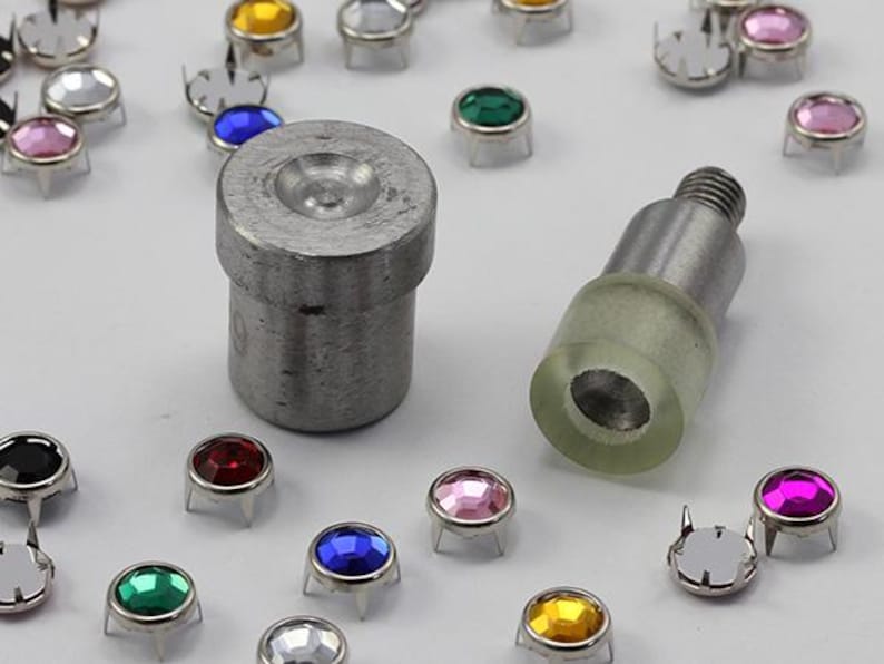 May include: An assortment of colorful rhinestone rivets and setting tools. The rivets have various colored gems, including pink, blue, green, and yellow. The tools are made of metal and are used for setting the rivets.