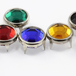 May include: Close-up of several colorful, faceted gemstone studs with silver metal settings. The gems are in various colors, including green, red, blue, yellow, black, and pink. Each stud has three metal prongs.