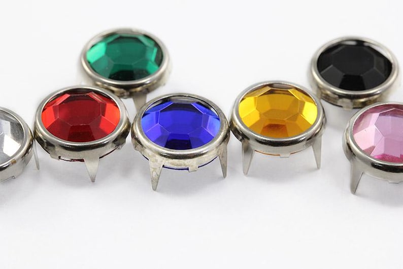 May include: Close-up of several colorful, faceted gemstone studs with silver metal settings. The gems are in various colors, including green, red, blue, yellow, black, and pink. Each stud has three metal prongs.