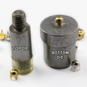 May include: Close-up of metal die tools for attaching studs. The top die has a threaded end, and the bottom die is cylindrical. The text "TOP DIE" and "BOTTOM DIE" are visible. Several gold and silver studs are scattered around the tools.