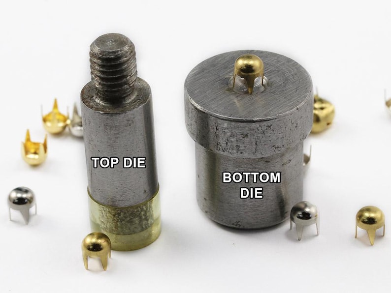 May include: Close-up of metal die tools for attaching studs. The top die has a threaded end, and the bottom die is cylindrical. The text "TOP DIE" and "BOTTOM DIE" are visible. Several gold and silver studs are scattered around the tools.