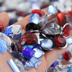 Puede incluir: Un puñado de gemas variadas en diversas formas y colores, incluyendo rojo, azul, transparente y negro. Las gemas están facetadas y reflejan la luz, con formas como corazones, estrellas y lágrimas. Ideal para la creación de joyas o proyectos de artesanía.