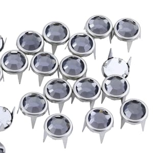 May include: A collection of silver-colored metal studs with gray, faceted, round gems. Each stud has three prongs. The gems have a reflective quality, and the studs are scattered against a white background.
