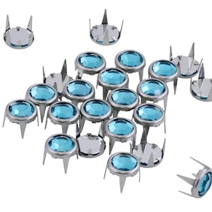 May include: A collection of silver-colored metal studs with turquoise-colored, oval-shaped gems. The studs have three prongs and are scattered across a white background. These decorative studs are suitable for various craft projects.