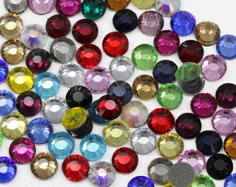 May include: Assortment of colourful round rhinestones in various shades of blue, green, red, yellow, purple, and clear. The rhinestones are scattered on a white surface.