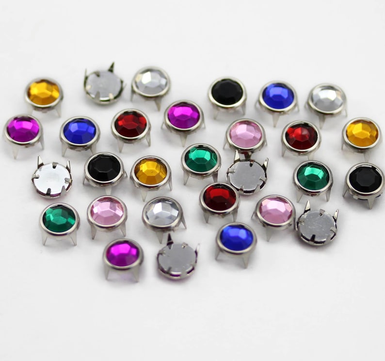 May include: A collection of colorful, round metal studs with faceted, jewel-like centers. The studs are in various colors, including yellow, pink, blue, red, green, and silver, and are arranged on a white surface. Each stud has a silver base with four prongs.