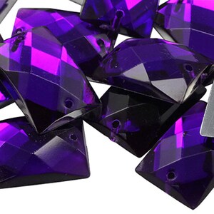 May include: A collection of faceted, rectangular purple crystal beads. Each bead has a hole for stringing and a reflective surface. The crystals are a deep violet color, with sharp angles and a glossy finish, suitable for jewelry making.