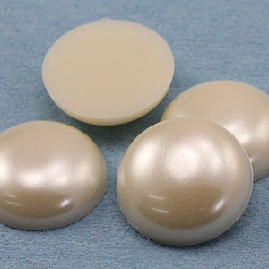 May include: Four ivory-colored, half-dome shaped craft beads. Three have a pearlescent finish, while one is a solid, matte ivory. The beads are arranged on a light blue textured surface.