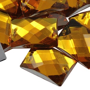 May include: Close-up of several faceted, amber-coloured glass rhinestones. The rhinestones are various geometric shapes, including squares and diamond shapes. Each has two small holes for attaching to fabric or other surfaces.