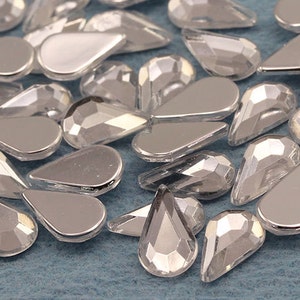 May include: A collection of silver-colored, teardrop-shaped rhinestones. Some rhinestones have a flat, reflective surface, while others have a faceted, multi-dimensional appearance. The rhinestones are scattered on a light blue surface.