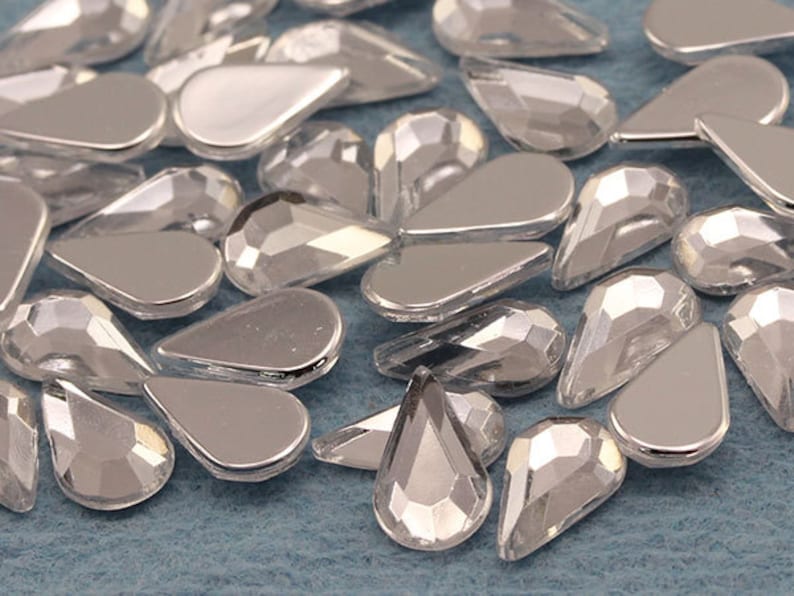 May include: A collection of silver-colored, teardrop-shaped rhinestones. Some rhinestones have a flat, reflective surface, while others have a faceted, multi-dimensional appearance. The rhinestones are scattered on a light blue surface.