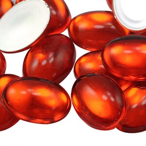 May include: A collection of oval, translucent red glass gems. One gem is white with a silver backing. The gems are scattered, with light reflecting off their smooth surfaces. Suitable for decorative use.