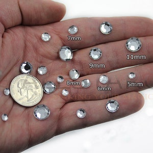 May include: A variety of clear, faceted rhinestones in different sizes, ranging from 5mm to 11mm, are displayed. A US quarter is included for size comparison. The rhinestones are held in a hand, showcasing their sparkle and clarity.