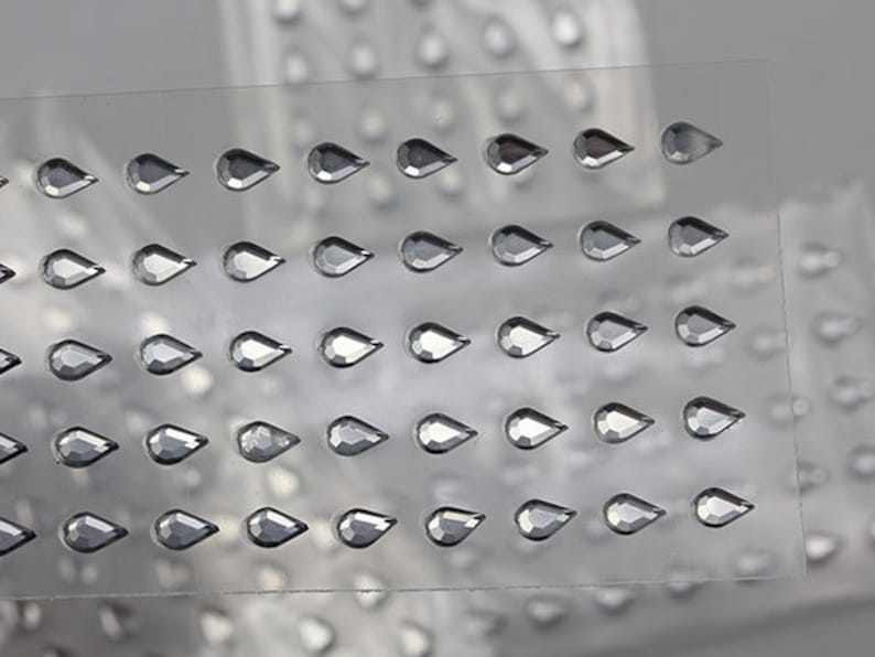 May include: Rows of clear, teardrop-shaped rhinestones are arranged on a clear, flexible sheet. The rhinestones have a faceted surface, reflecting light. The image shows multiple sheets of these rhinestones, suggesting a craft or embellishment product.
