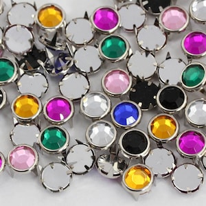 May include: A collection of colorful rhinestone studs with silver metal bases. The rhinestones are in various colors, including pink, green, yellow, and black. The studs have a pointed back.