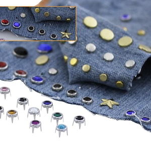May include: Close-up of denim fabric embellished with various metal studs and colorful gemstones. The studs are in gold, silver, and black, while the gemstones are red, blue, and purple. A gold star-shaped stud is also visible.
