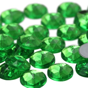 Peut inclure: Un grand nombre de petits joyaux ronds, verts, facetés, en verre. Les joyaux sont éparpillés sur une surface blanche.