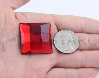 May include: A red square-shaped crystal rhinestone with a faceted design, next to a US quarter for size comparison.