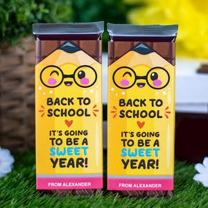 Op de afbeelding: Twee chocoladerepen met gele verpakkingen met een potloodthema. De verpakkingen bevatten de tekst "Back to School" en "It's going to be a sweet year!" met een schattig potloodpersonage met een bril. De onderkant van de verpakking zegt "From Alexander".