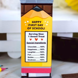 Op de afbeelding: Een rechthoekige snoepreepverpakking met een potloodontwerp en de tekst "HAPPY FIRST DAY OF SCHOOL!". De verpakking vermeldt "Speciale Ingredi&euml;nten" waaronder chocolade, geluk, lachen, plezier, glimlachen en vriendelijkheid.