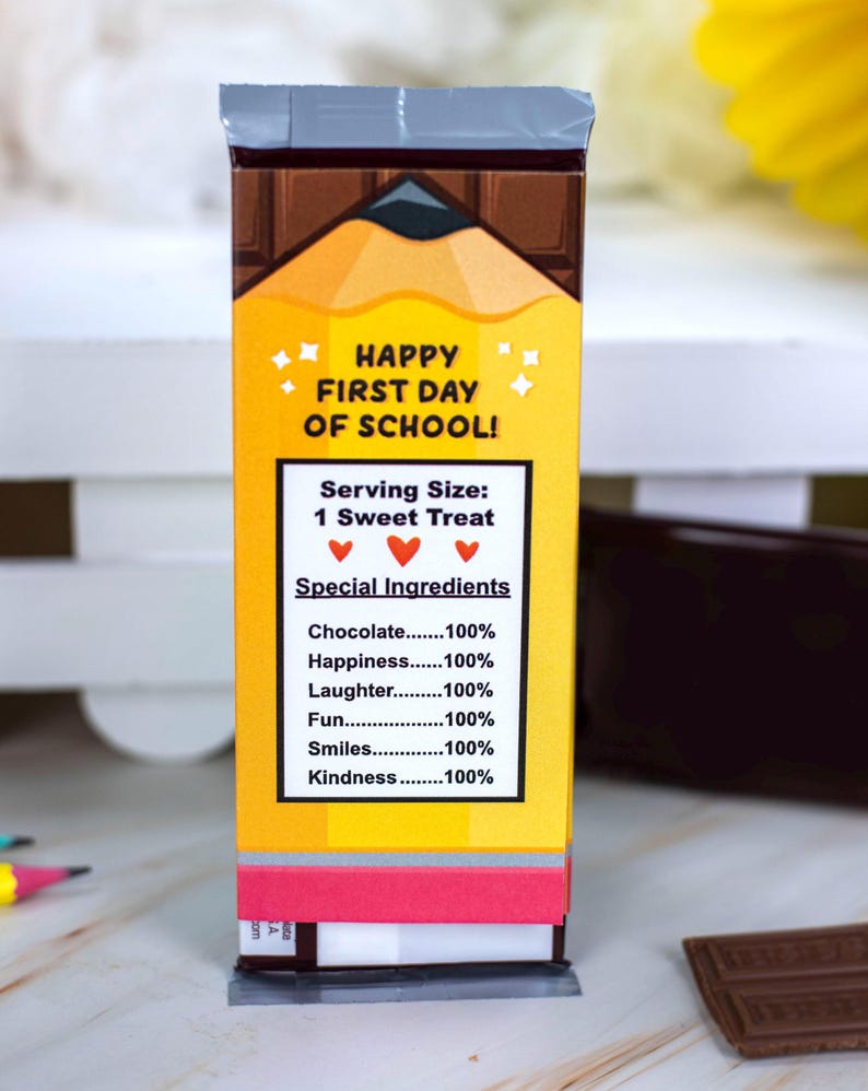 Op de afbeelding: Een rechthoekige snoepreepverpakking met een potloodontwerp en de tekst "HAPPY FIRST DAY OF SCHOOL!". De verpakking vermeldt "Speciale Ingredi&euml;nten" waaronder chocolade, geluk, lachen, plezier, glimlachen en vriendelijkheid.