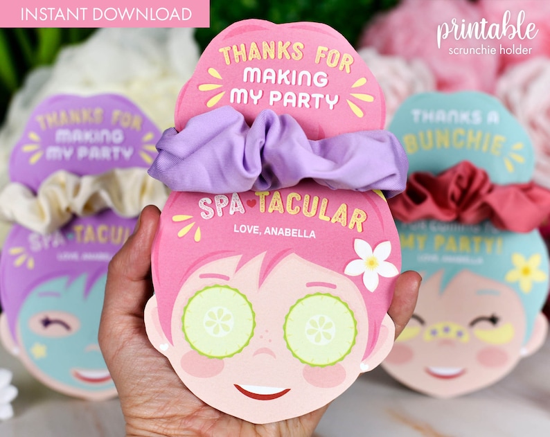 K&ouml;nnte beinhalten: Ein rosa Scrunchie-Halter mit Spa-Thema. Der Halter zeigt ein Cartoon-Gesicht mit Gurkenscheiben auf den Augen und den Text "SPA-TACULAR" und "THANKS FOR MAKING MY PARTY". Ein lila Scrunchie ist befestigt.