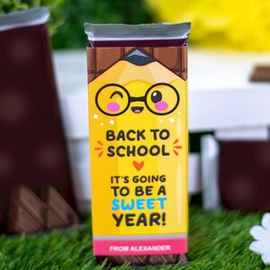 Op de afbeelding: Een chocoladereep met een gele verpakking met een potloodontwerp en de tekst "BACK TO SCHOOL". De verpakking zegt ook "IT'S GOING TO BE A SWEET YEAR!" en "FROM ALEXANDER". De verpakking heeft een cartoon potlood met een bril.