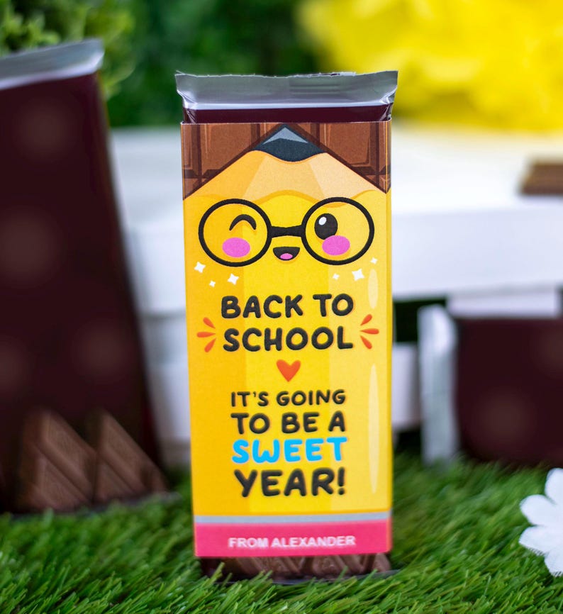 Op de afbeelding: Een chocoladereep met een gele verpakking met een potloodontwerp en de tekst "BACK TO SCHOOL". De verpakking zegt ook "IT'S GOING TO BE A SWEET YEAR!" en "FROM ALEXANDER". De verpakking heeft een cartoon potlood met een bril.