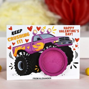 May include: A Valentine's Day card featuring a cartoon monster truck with the text "Keep Crushing It!" and "Happy Valentine's Day!"  The card includes a pink play dough container with the word "Play-Doh" printed on it. The card is signed "From Alexander."