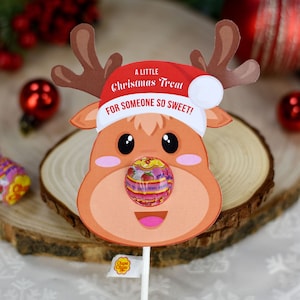 Puede incluir: Un soporte de piruleta imprimible con forma de cabeza de reno con el texto "A Little Christmas Treat For Someone So Sweet!" impreso en &eacute;l. El reno lleva un gorro de Pap&aacute; Noel rojo y una piruleta insertada en su nariz.