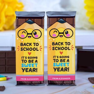 Op de afbeelding: Twee chocoladerepen met gele en roze etiketten. De etiketten tonen een cartoonpotlood met een bril en de tekst "Back to School, It's Going to Be a Sweet Year!" en "From Alexander."