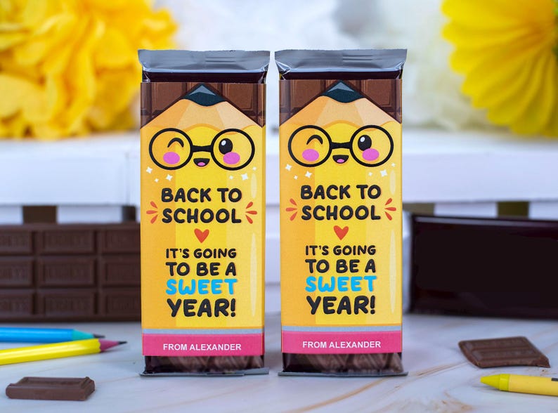 Op de afbeelding: Twee chocoladerepen met gele en roze etiketten. De etiketten tonen een cartoonpotlood met een bril en de tekst "Back to School, It's Going to Be a Sweet Year!" en "From Alexander."