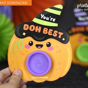 May include: Printable Halloween-themed play dough holder shaped like a pumpkin with a witch hat. The pumpkin has a cute face and the text "You're DOH BEST" on it. The holder has a purple play dough container in the center.