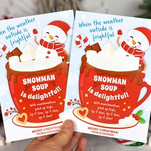 Puede incluir: Dos tarjetas de Navidad con un mu&ntilde;eco de nieve en una taza roja de chocolate caliente con malvaviscos. El texto en las tarjetas dice "Cuando el clima afuera es espantoso, &iexcl;la sopa de mu&ntilde;eco de nieve es deliciosa!" con malvaviscos apilados en alto, &iexcl;b&eacute;belo despacio, b&eacute;belo despacio, b&eacute;belo despacio! &iexcl;Feliz Navidad! De parte de Alexander.