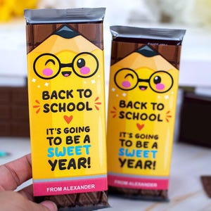 Op de afbeelding: Twee chocoladerepen met gele verpakkingen met een potloodontwerp en de tekst "Back to School". De verpakkingen zeggen ook "It's going to be a sweet year!" en "From Alexander".