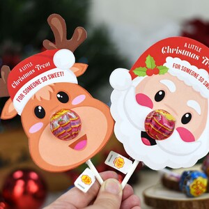 Puede incluir: Dos soportes para piruletas con tem&aacute;tica navide&ntilde;a, uno con forma de reno y el otro con forma de Pap&aacute; Noel. Ambos tienen una piruleta pegada a la nariz, y el texto "A Little Christmas Treat For Someone So Sweet!" est&aacute; impreso en cada uno.