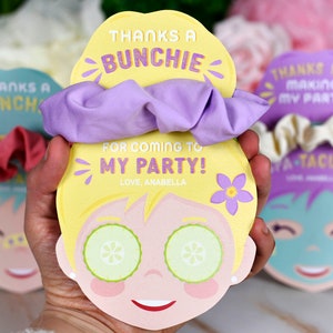 K&ouml;nnte beinhalten: Ein Party-Geschenkset mit einer gelben Karte in Form eines Gesichts, Gurkenscheiben, einem lila Scrunchie und dem Text: "THANKS A BUNCHIE FOR COMING TO MY PARTY! LOVE, ANABELLA". Weitere Karten mit Scrunchies sind sichtbar.