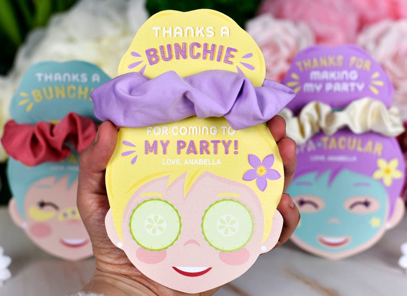K&ouml;nnte beinhalten: Ein Party-Geschenkset mit einer gelben Karte in Form eines Gesichts, Gurkenscheiben, einem lila Scrunchie und dem Text: "THANKS A BUNCHIE FOR COMING TO MY PARTY! LOVE, ANABELLA". Weitere Karten mit Scrunchies sind sichtbar.