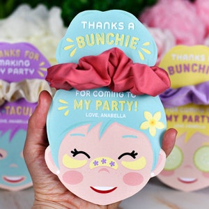 K&ouml;nnte beinhalten: Party-Gunstkarten mit Scrunchies. Die Karten sind wie ein Gesicht geformt und tragen den Text "THANKS A BUNCHIE FOR COMING TO MY PARTY! LOVE, ANABELLA". Die Scrunchies sind rosa, lila und cremefarben.