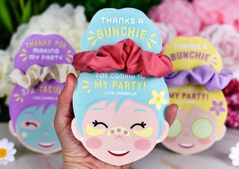 K&ouml;nnte beinhalten: Party-Gunstkarten mit Scrunchies. Die Karten sind wie ein Gesicht geformt und tragen den Text "THANKS A BUNCHIE FOR COMING TO MY PARTY! LOVE, ANABELLA". Die Scrunchies sind rosa, lila und cremefarben.