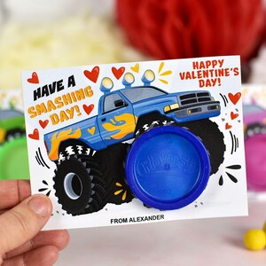 May include: A blue monster truck with yellow flames on the side, a blue Play-Doh container, and a Valentine's Day card that says "Have a Smashing Day!" and "Happy Valentine's Day!" The card is signed "From Alexander."
