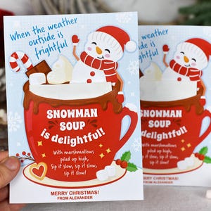 Puede incluir: Una tarjeta de Navidad con una taza roja llena de chocolate caliente y malvaviscos, coronada por un mu&ntilde;eco de nieve. La tarjeta dice "Snowman Soup is delightful!" con el texto "Merry Christmas! From Alexander" en la parte inferior.