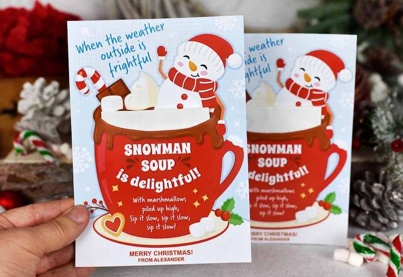 Puede incluir: Una tarjeta de Navidad con una taza roja llena de chocolate caliente y malvaviscos, coronada por un mu&ntilde;eco de nieve. La tarjeta dice "Snowman Soup is delightful!" con el texto "Merry Christmas! From Alexander" en la parte inferior.