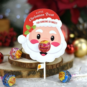 Puede incluir: Un recorte de cara de Pap&aacute; Noel blanco con un gorro rojo y una piruleta pegada en la nariz. La piruleta es roja y rosa con las palabras "Chupa Chups Fresa" impresas en ella. El recorte tiene el texto "&iexcl;Un peque&ntilde;o regalo de Navidad para alguien tan dulce!" impreso en &eacute;l.