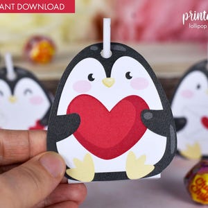 May include: Printable penguin lollipop holder with a red heart. The penguin is black and white with a yellow beak and feet. The holder is designed to be used with a lollipop.