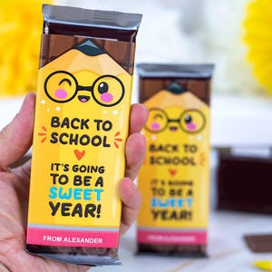 Op de afbeelding: Gele chocoladereepverpakking met een potloodontwerp en de tekst "BACK TO SCHOOL, IT'S GOING TO BE A SWEET YEAR!". De verpakking heeft een cartoonpotlood met een bril. De onderkant van de verpakking luidt "FROM ALEXANDER".