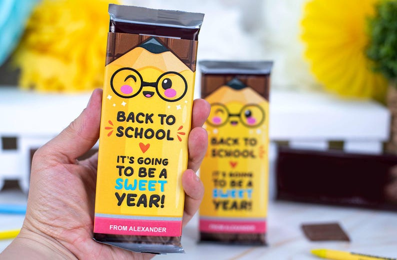 Op de afbeelding: Gele chocoladereepverpakking met een potloodontwerp en de tekst "BACK TO SCHOOL, IT'S GOING TO BE A SWEET YEAR!". De verpakking heeft een cartoonpotlood met een bril. De onderkant van de verpakking luidt "FROM ALEXANDER".